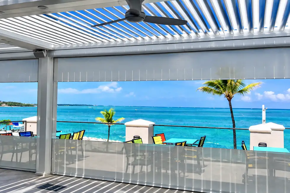 Motorized outdoor screens lowered around a white louvered pergola overlooking a bright waterfront setting